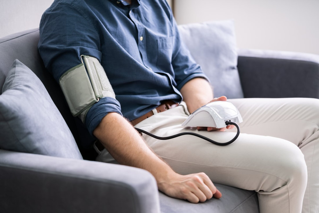 Ein Mann sitzt auf einer grauen Couch mit einer Manschette um den Arm und einem Blutdruckmessgerät und misst Blutdruck und Herzfrequenz