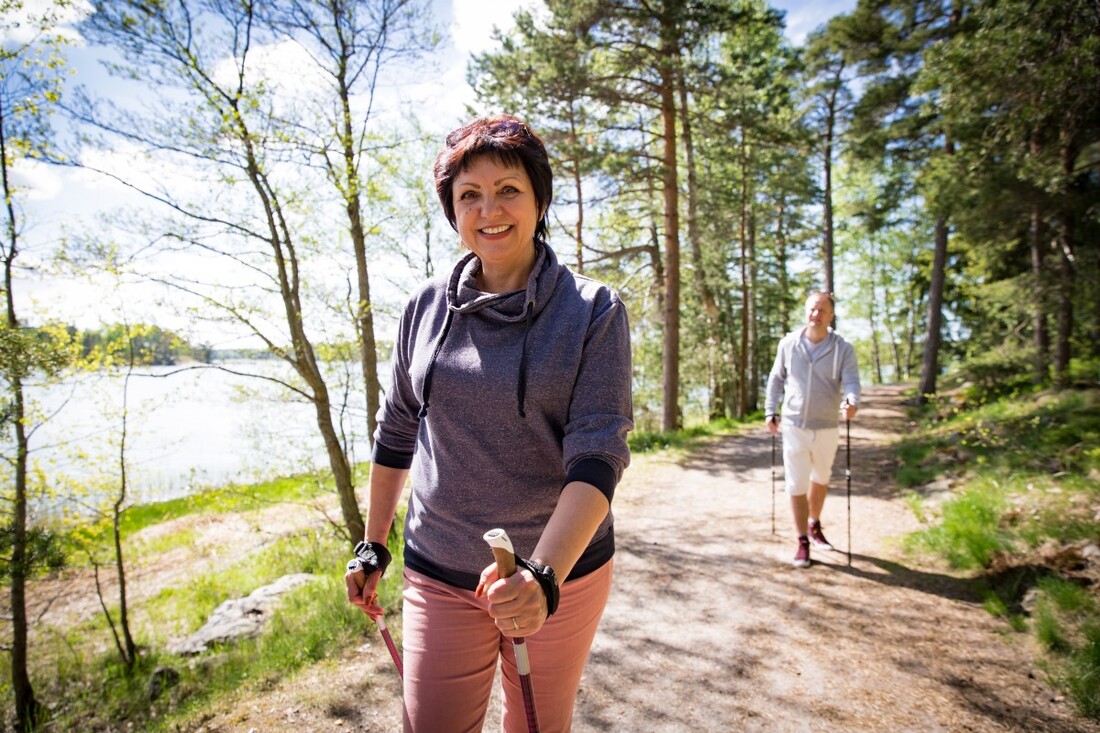 Eine Frau mittleren Alters läuft lächelnd mit Nordic Working Stöcken über einen sonnigen Waldweg